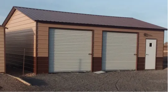 Two beige garage doors on a brown metal utility building with a white side door, on gravel under a clear sky