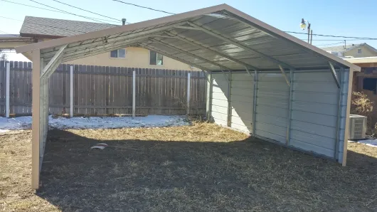 Open-sided metal carport with a slanted roof in a gravel yard, wooden fence in the background.