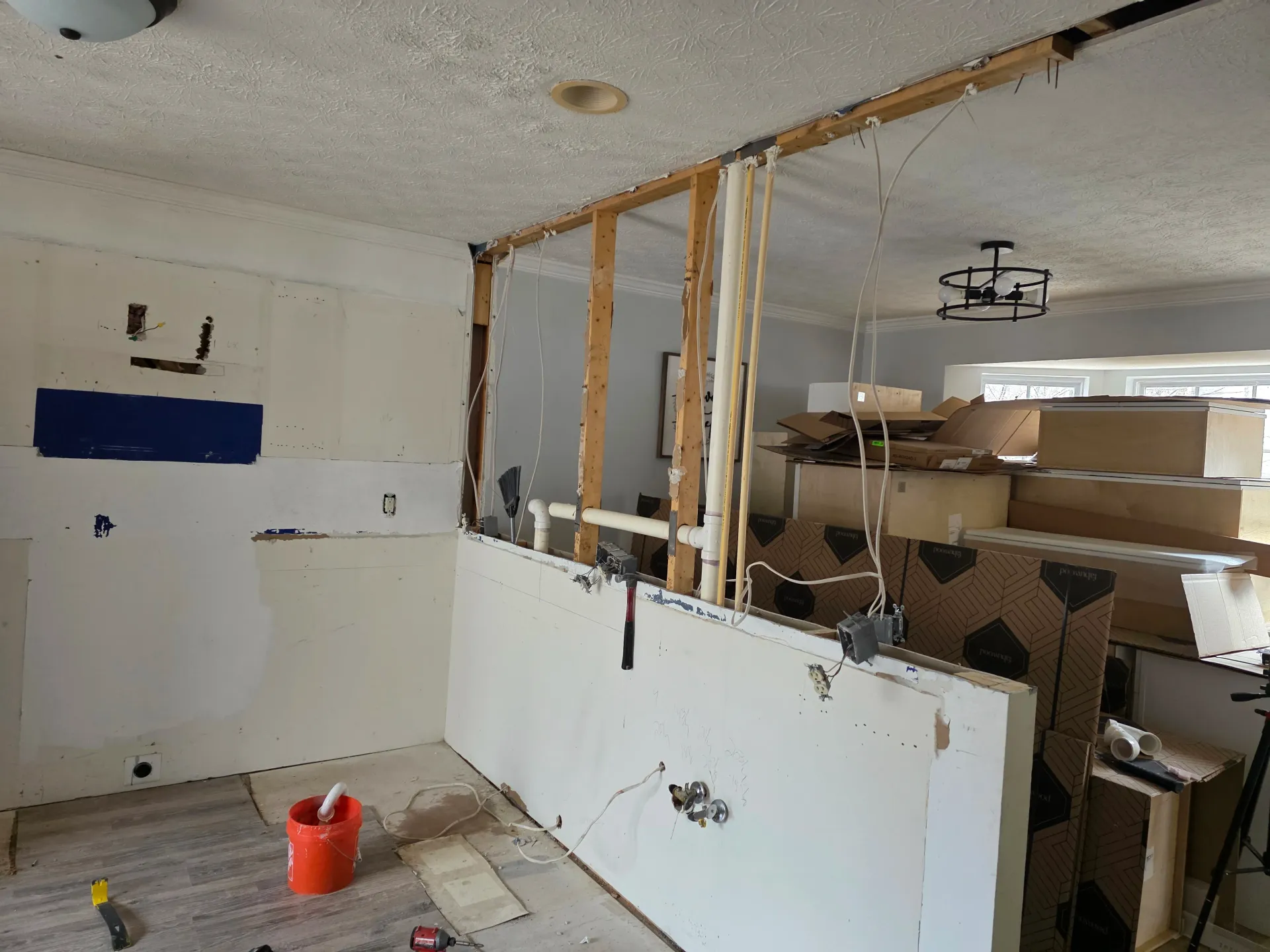 A room undergoing renovation with a partially demolished wall revealing wooden studs, exposed wiring, and cardboard boxes.