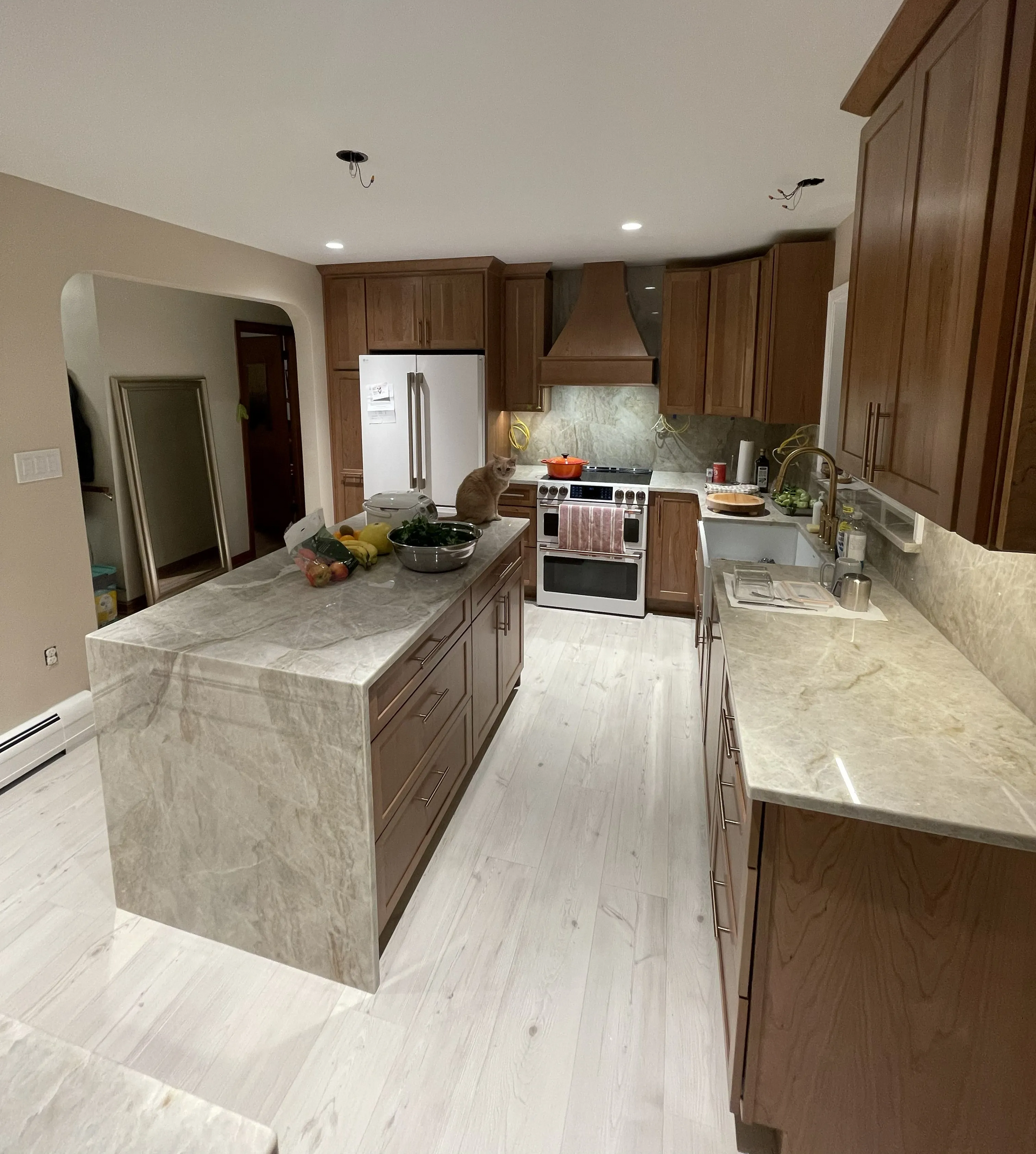 A modern kitchen featuring a large stone-topped island, wooden cabinetry, stainless steel appliances, and light wood floors.