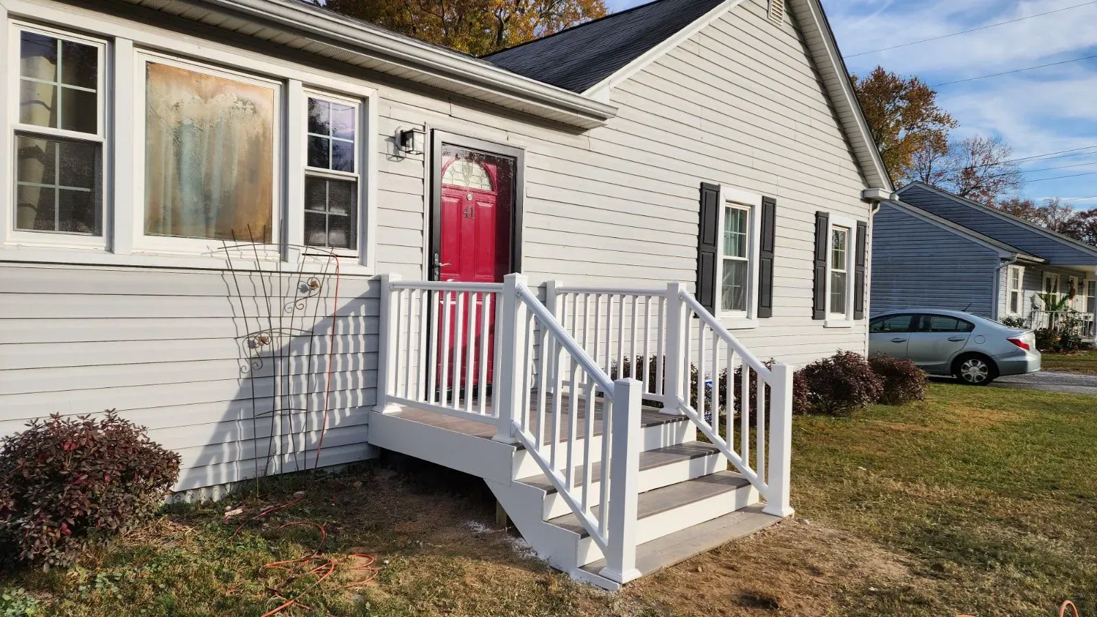 White house with a red front door, white railings, and front steps, surrounded by a yard under a blue sky.