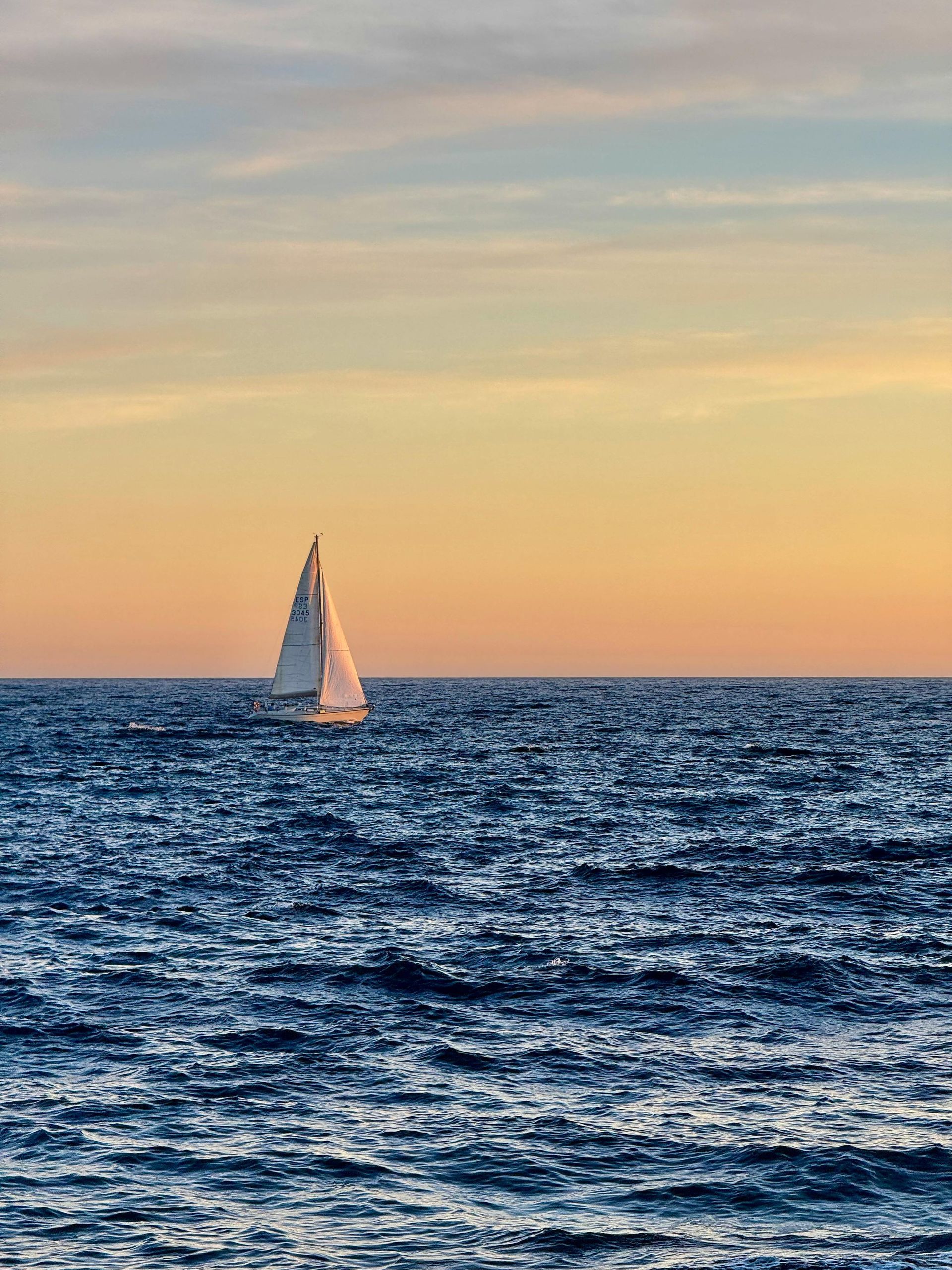 Sailboat on a choppy blue sea under a sunset of yellow and orange.