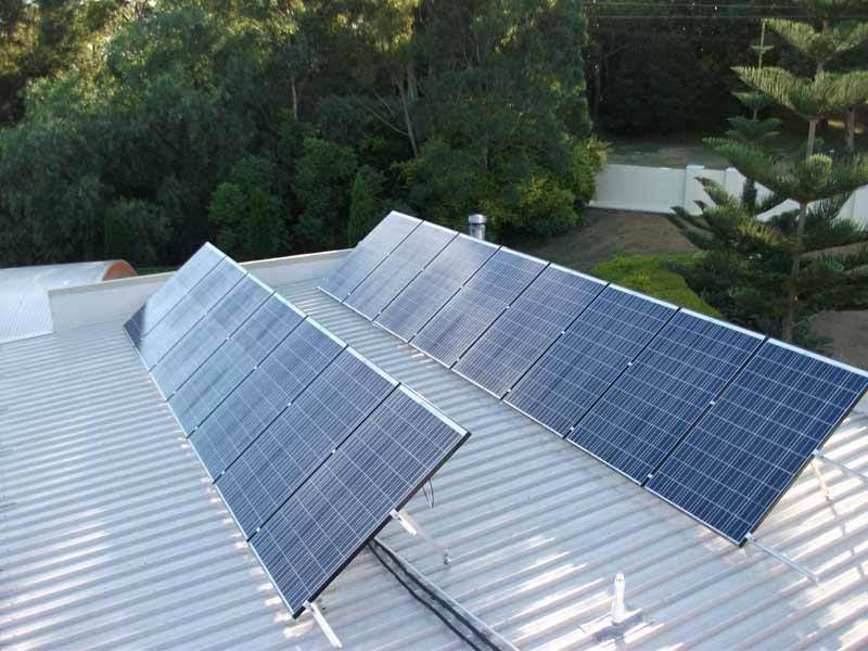 A Row Of Solar Panels On A Roof With Trees In The Background — CJ Electrical & Solar In Swansea, NSW