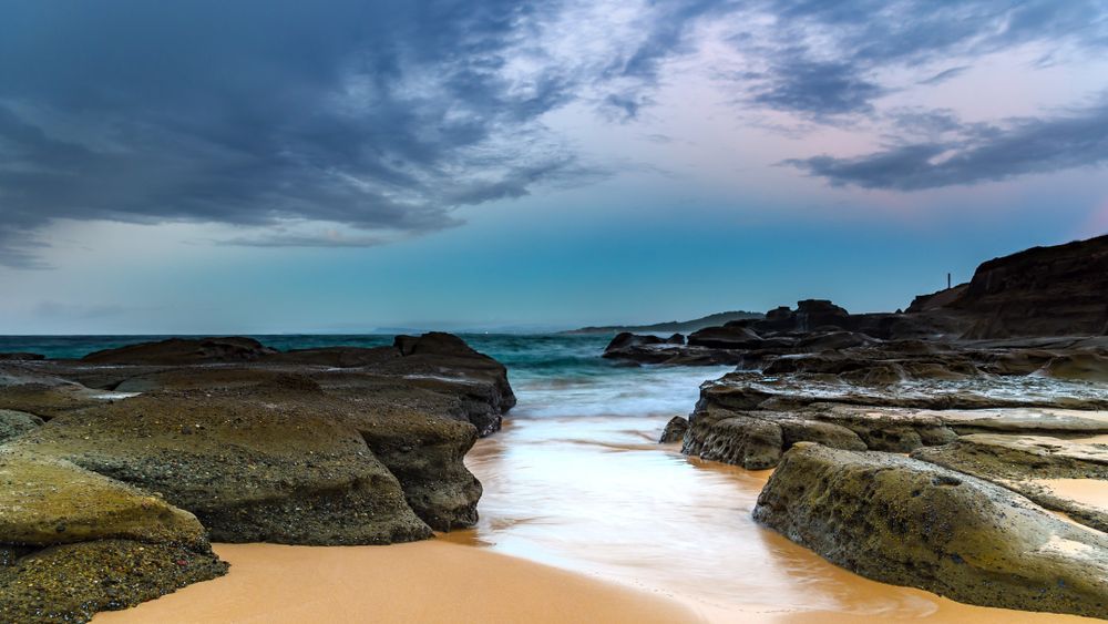 A Beach With Rocks And Waves Coming In On A Cloudy Day — CJ Electrical & Solar In Wyong, NSW