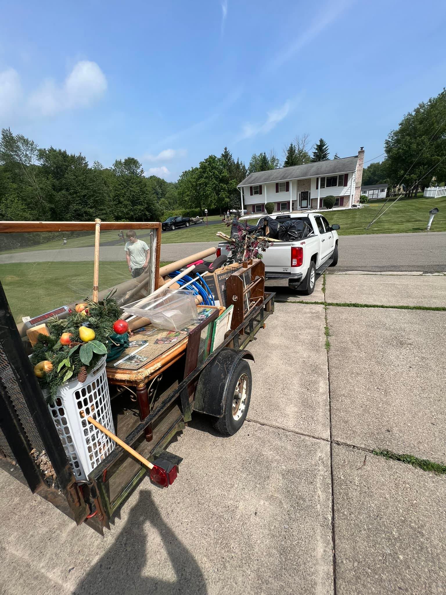 A white pickup truck pulls a trailer filled with debris in front of a house on a sunny day.