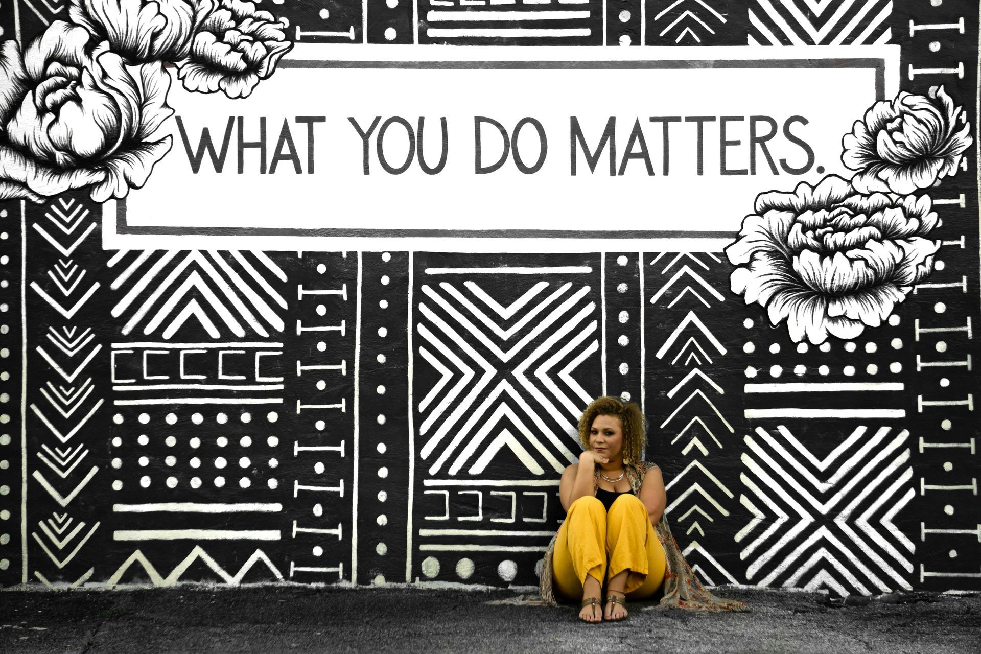 Woman in yellow trousers sitting on the floor against a black wall that has white geometric patterns
