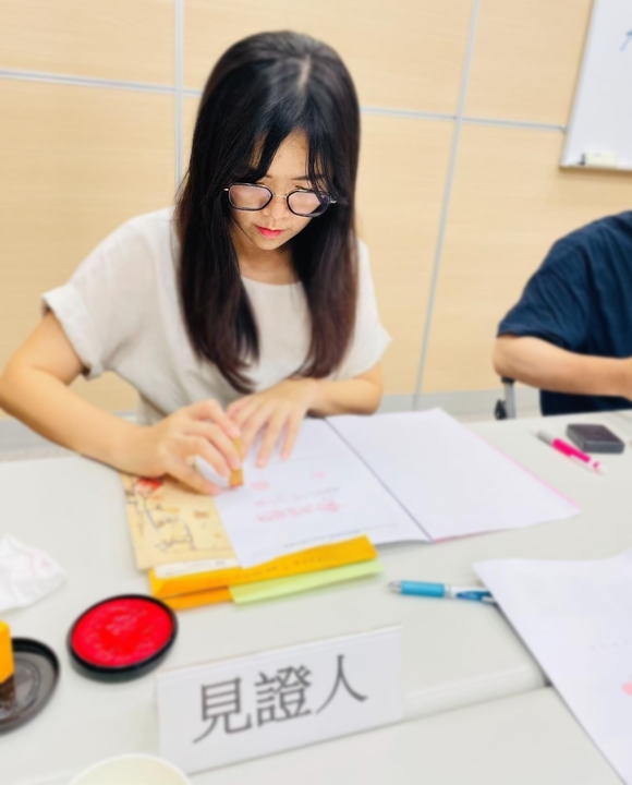 詠華地政士事務所所長－鄭秀慧女士在桌前蓋章。 「見證人」標誌清晰可見。桌上還有紅色印泥和筆記本。