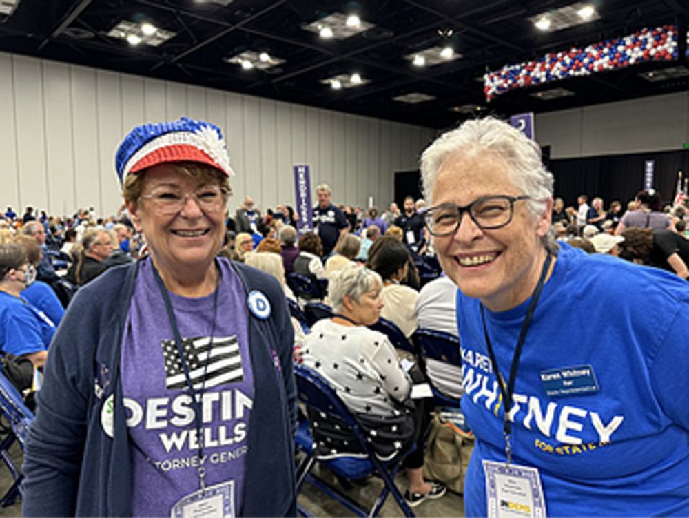 Two women smiling at a political event. One wears a hat, the other a blue shirt. People in background.