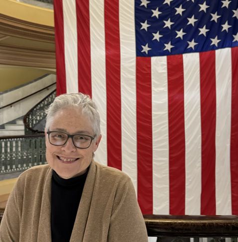 Woman smiles in front of a large American flag. She wears glasses, a tan cardigan, and a black shirt.