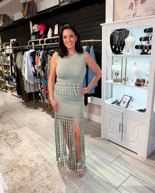 Woman in a light gray fringed dress standing in a boutique showroom, smiling with hand on hip.