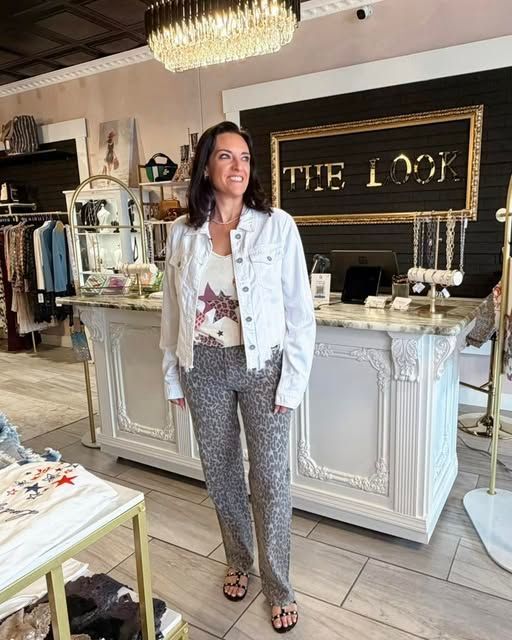 Woman standing in a boutique, wearing a light jacket and patterned pants, with “THE LOFT” sign behind her