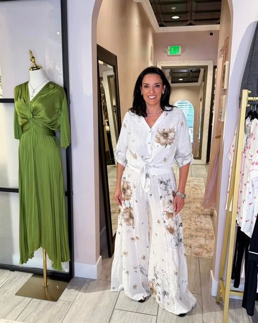 Woman in a white floral jumpsuit stands in a boutique hallway beside a green dress on a mannequin.
