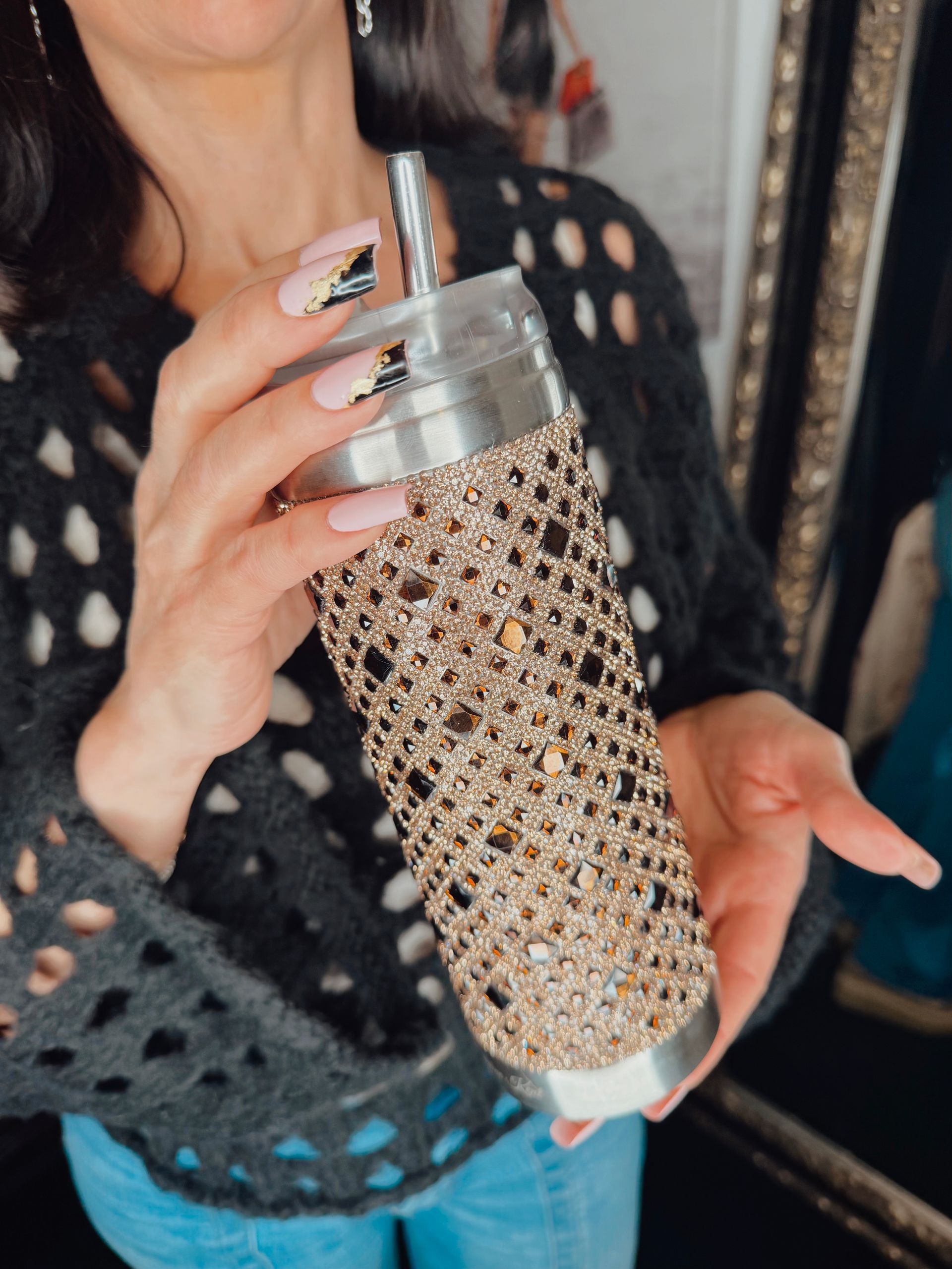 Person holding a rhinestone-covered tumbler with a straw, wearing a black top and blue jeans.