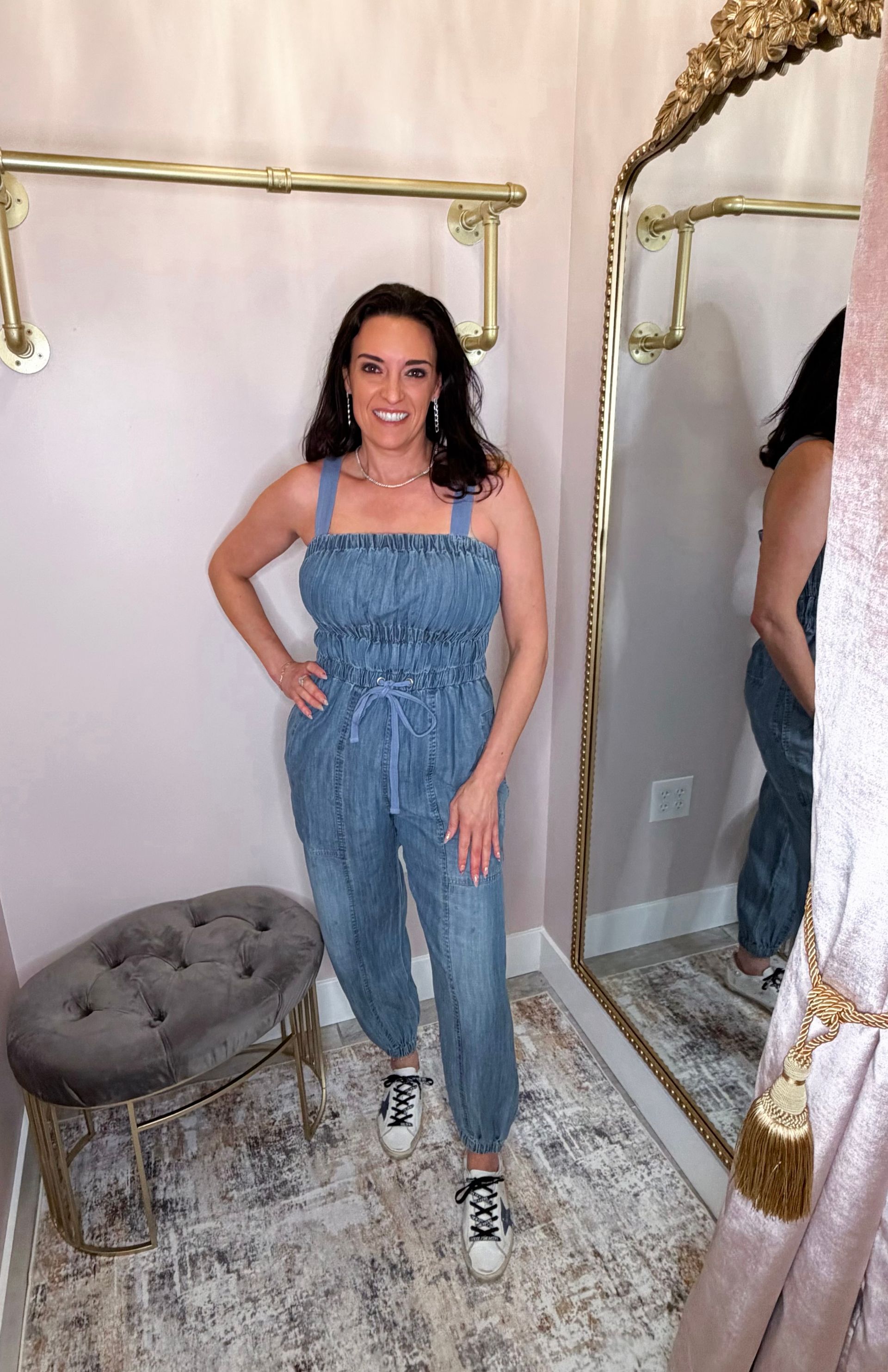 Woman standing in a mirrored room, wearing a blue jumpsuit and smiling.