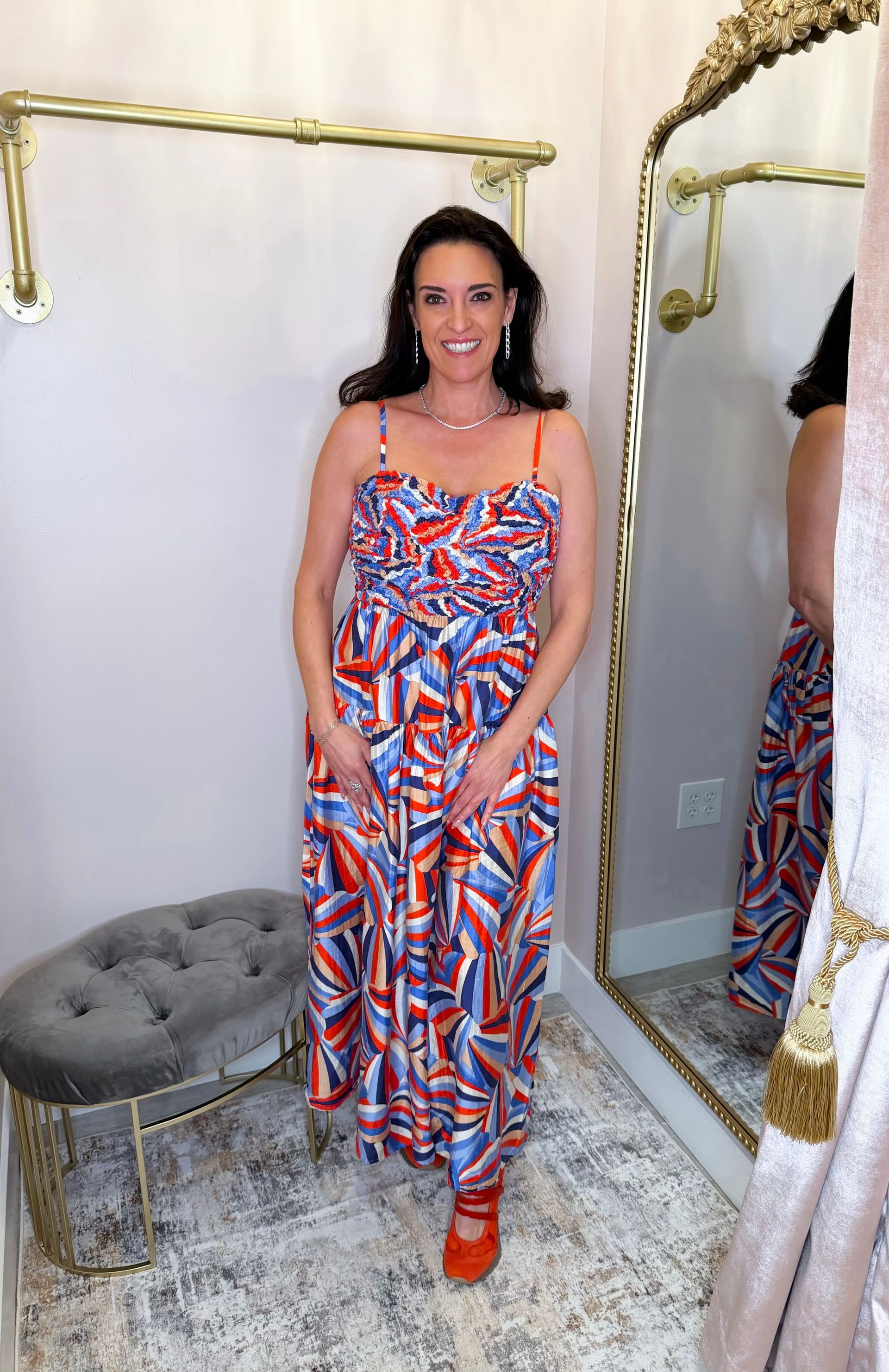 Woman in a colorful patterned dress stands in a boutique fitting room with a mirror and stool.