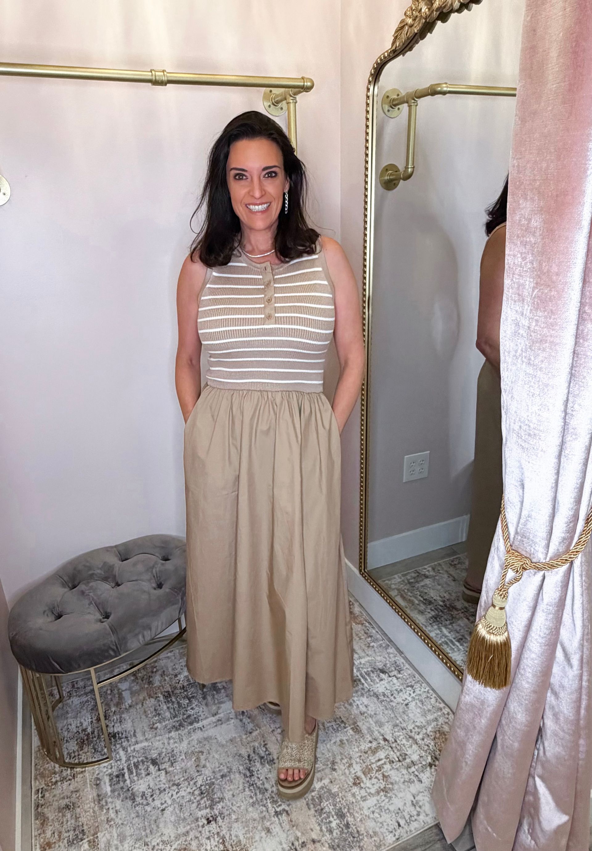 Woman in a beige dress posing in a dressing room with a mirror and tufted stool.
