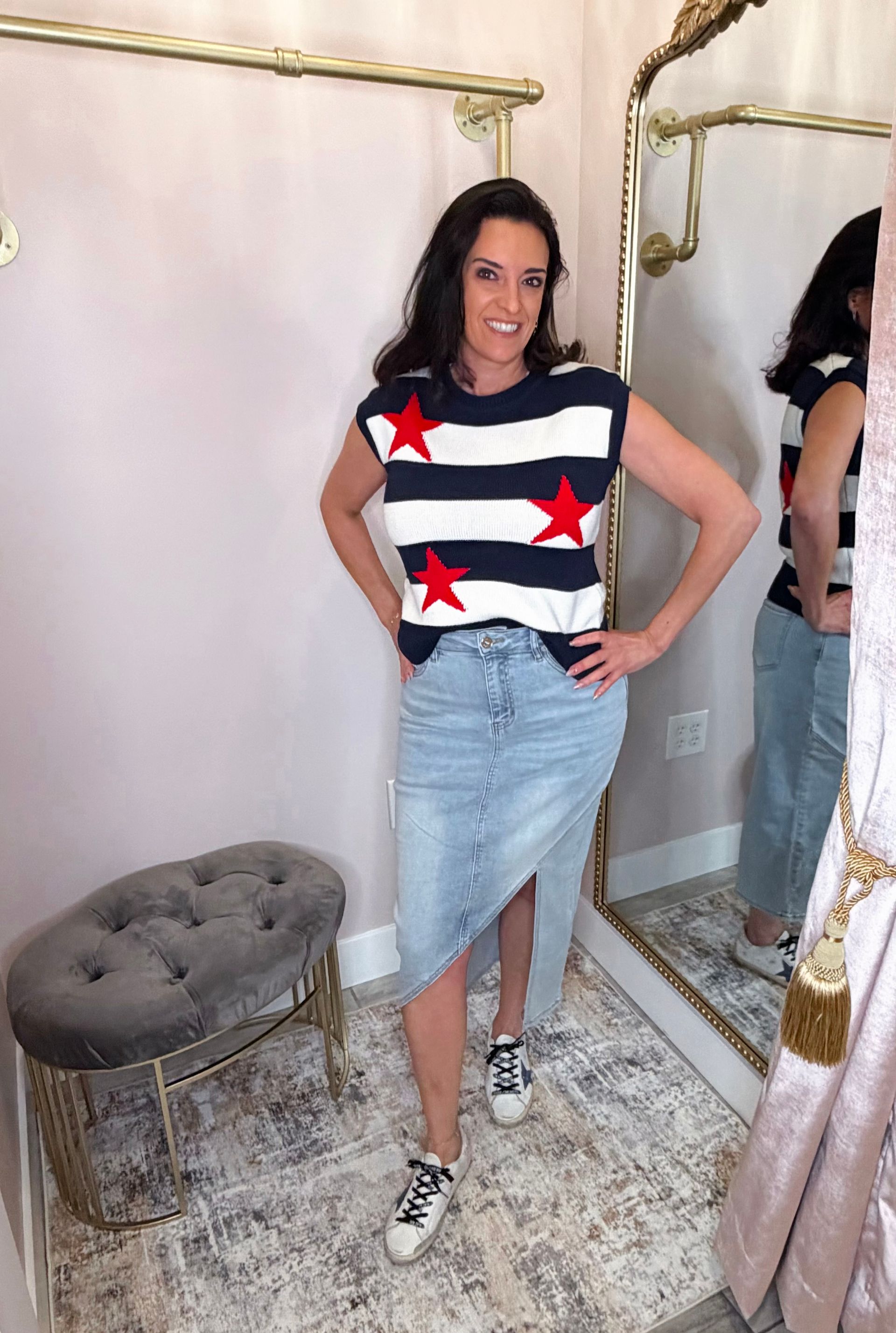 Woman posing in a mirror, wearing a star-patterned top, denim skirt, and sneakers in a boutique fitting room
