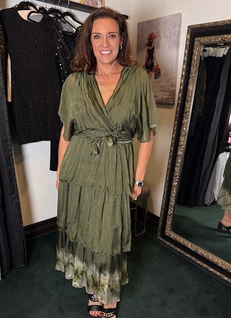 Woman standing in a boutique, wearing an olive-green dress beside a full-length mirror