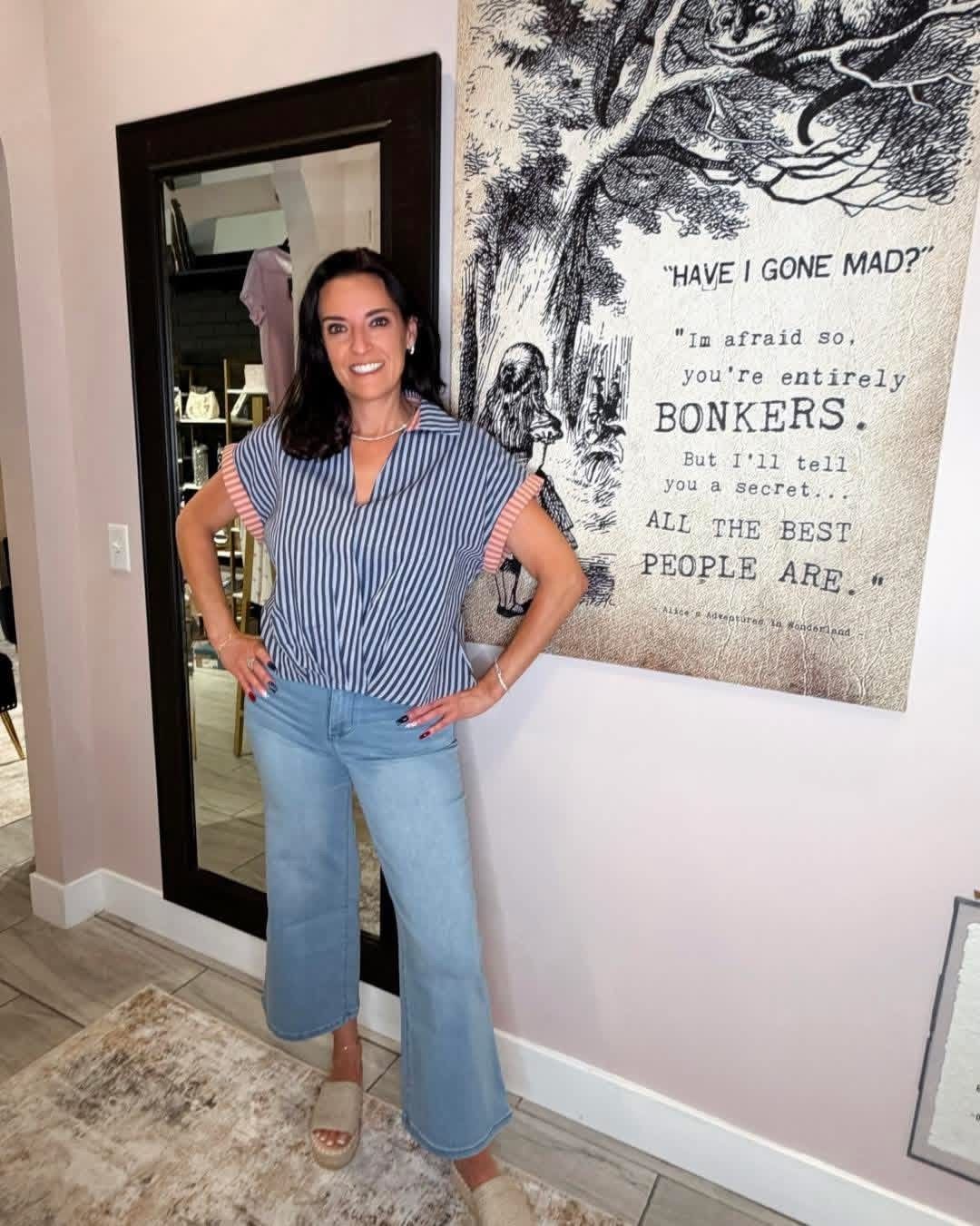 Woman standing indoors by a mirror and wall art, wearing a striped blouse and jeans.