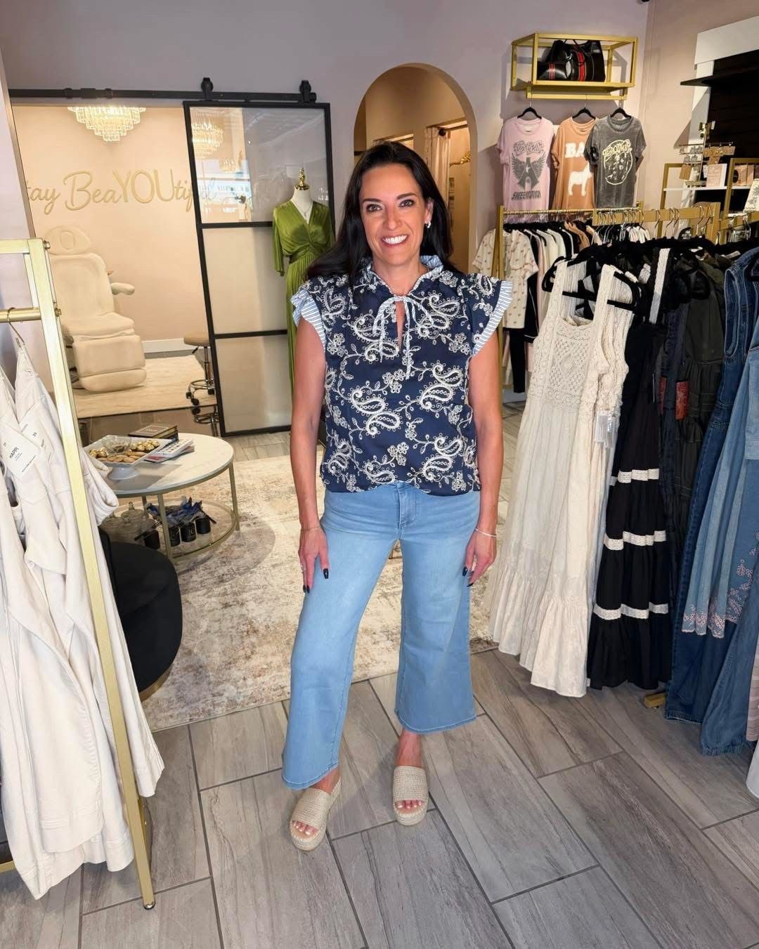 Woman smiling in a boutique, wearing a navy floral blouse, light jeans, and sandals.