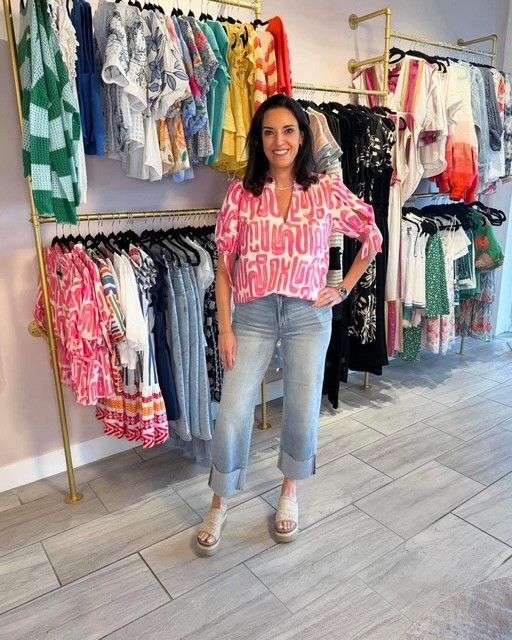 Woman standing in a clothing boutique, wearing a pink patterned top, jeans, and sandals.