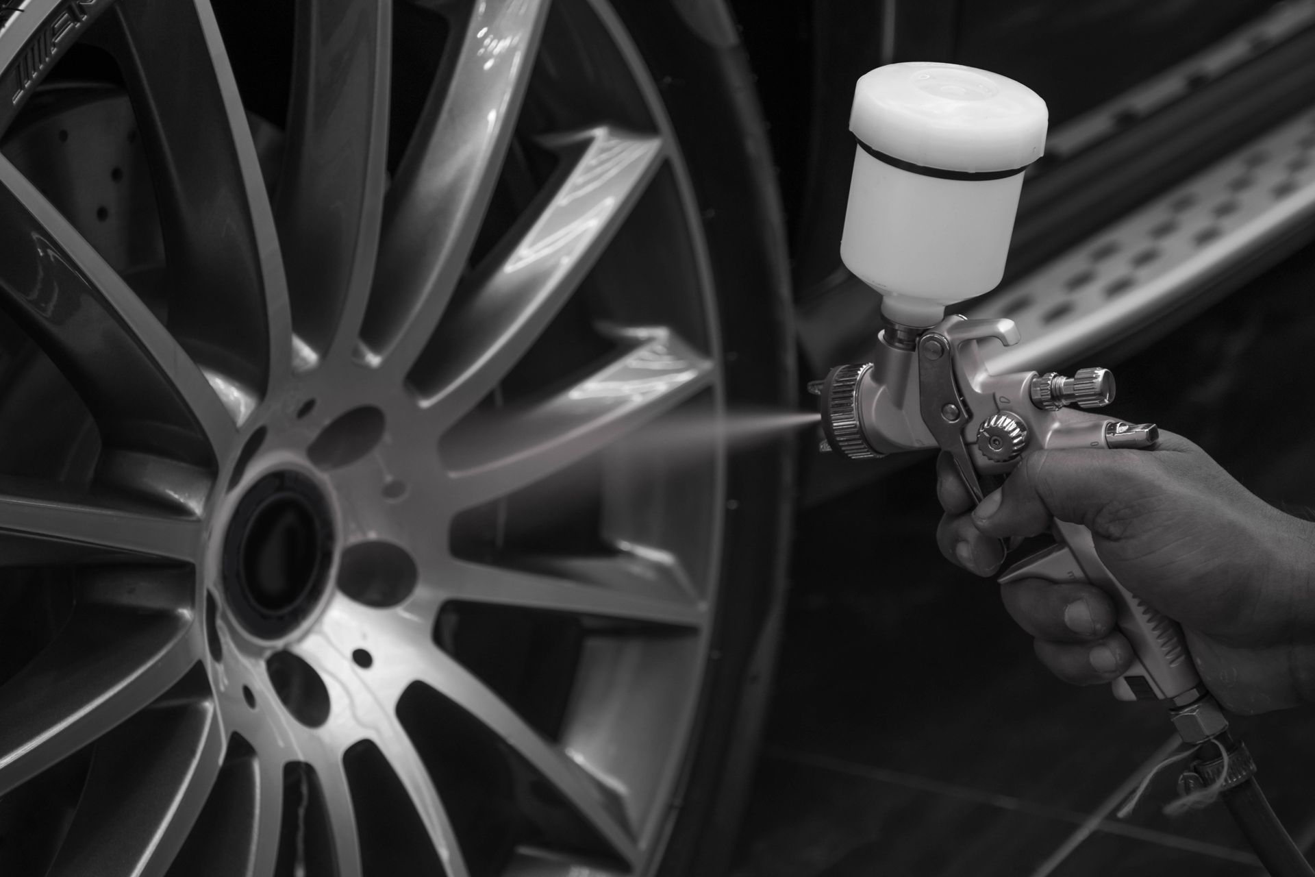 Spray gun painting a car wheel rim in a workshop, with paint mist visible.