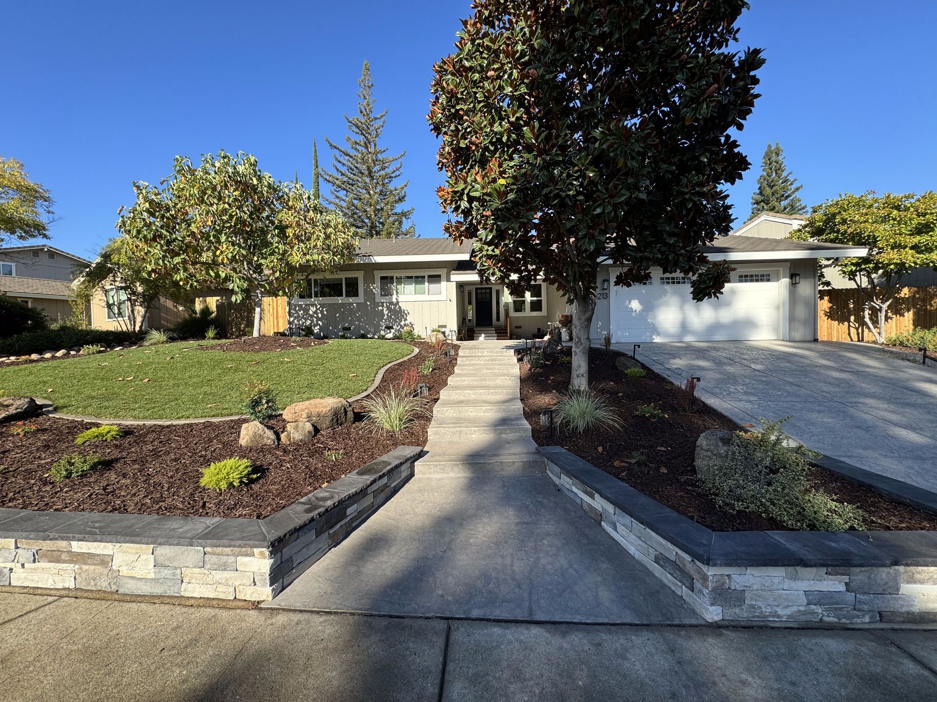 A house with a concrete walkway leading to it and a tree in front of it.