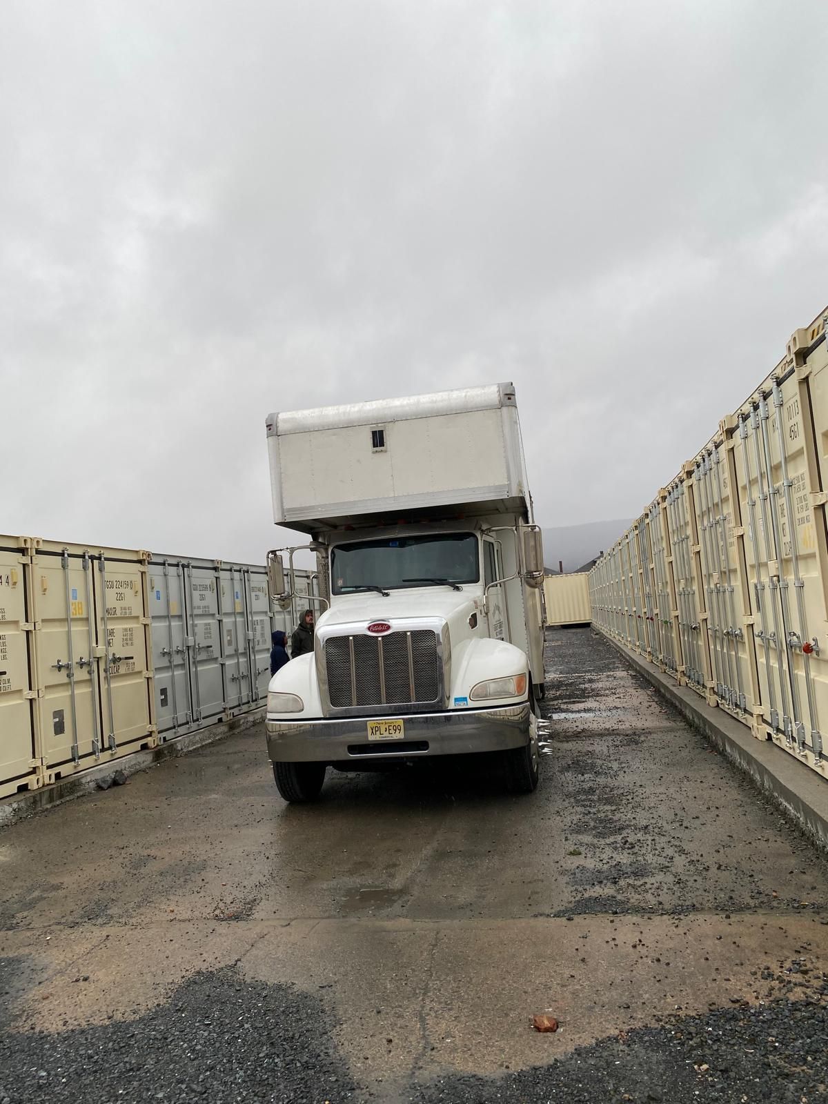 A white truck is parked in a parking lot next to a row of shipping containers.