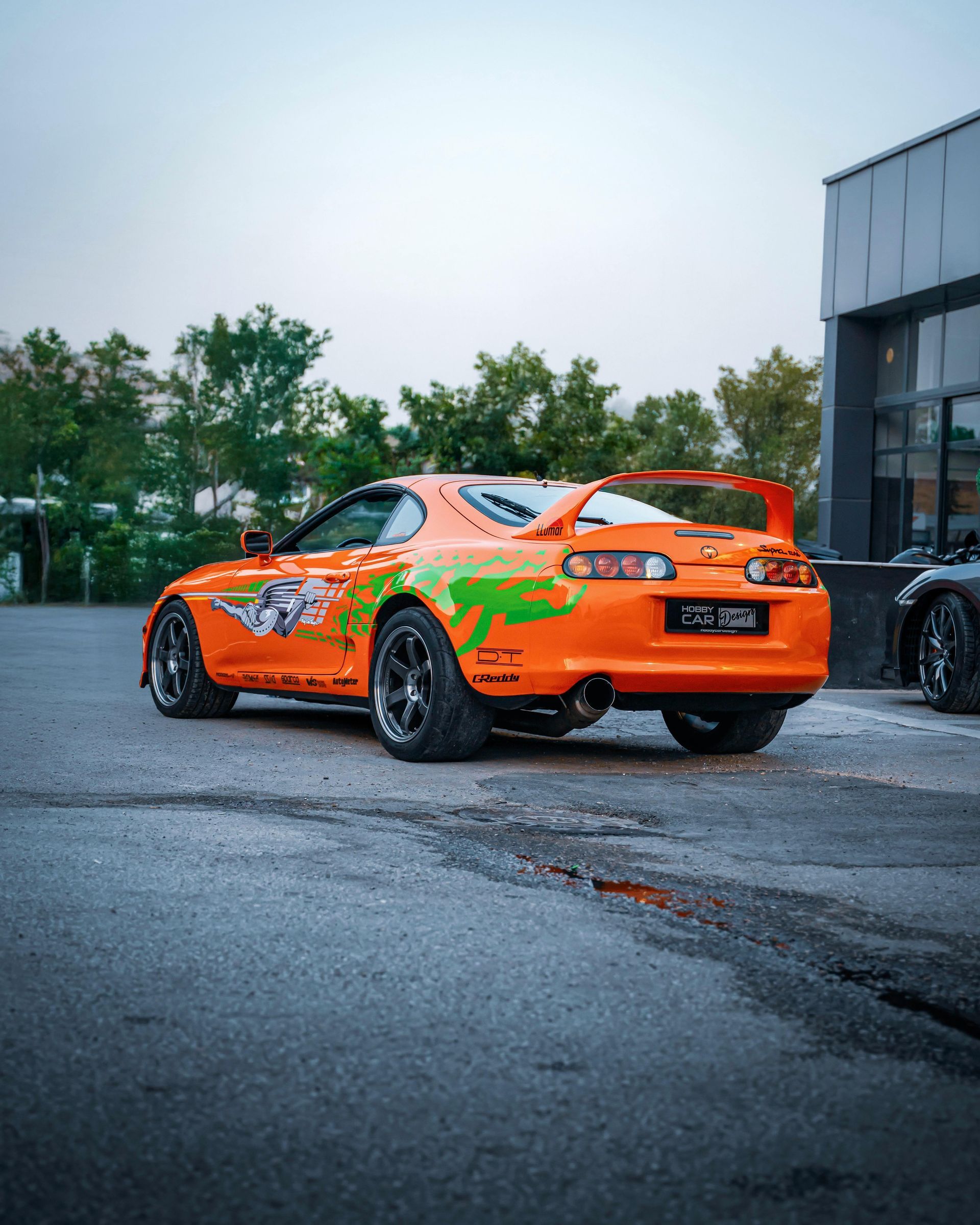 Orange sports car with green graphics parked on wet pavement beside a building