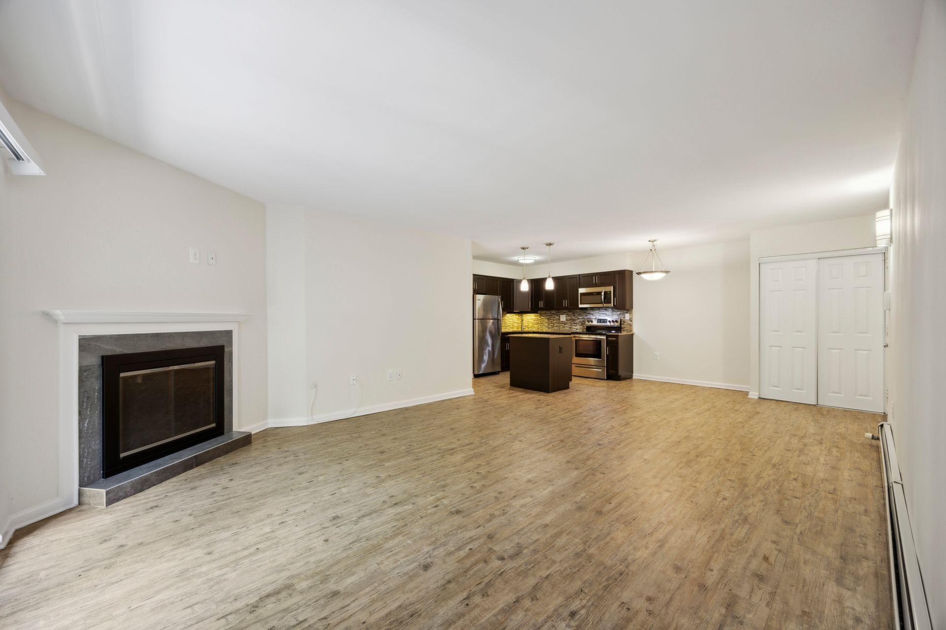 Empty living room with fireplace, kitchen in the background, light-colored flooring and walls.