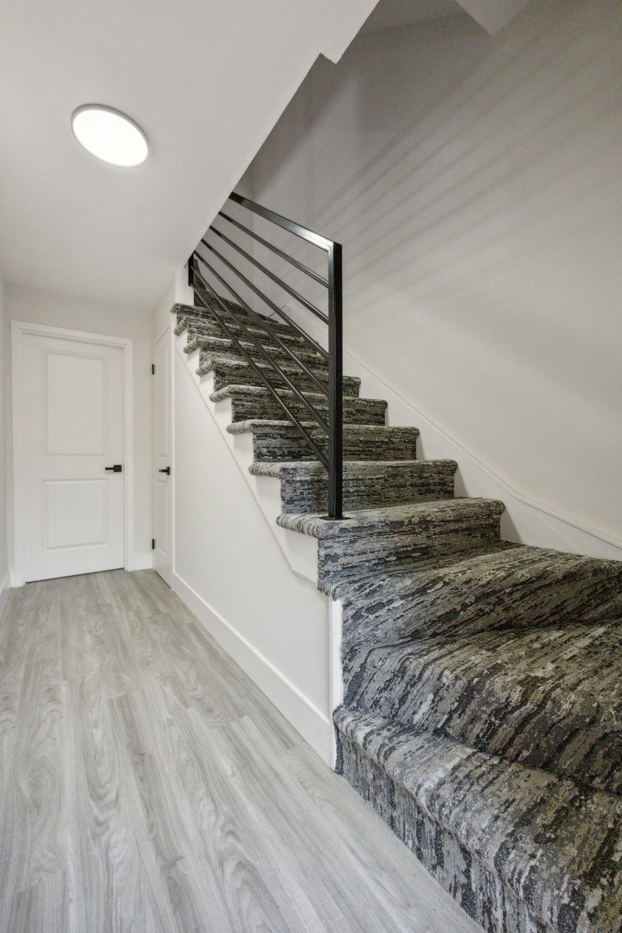Staircase with gray patterned carpet, black railing, white walls, door, and light wood-look floor.