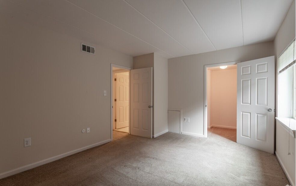 Empty bedroom with beige walls, carpet, and three white doors leading to other rooms.