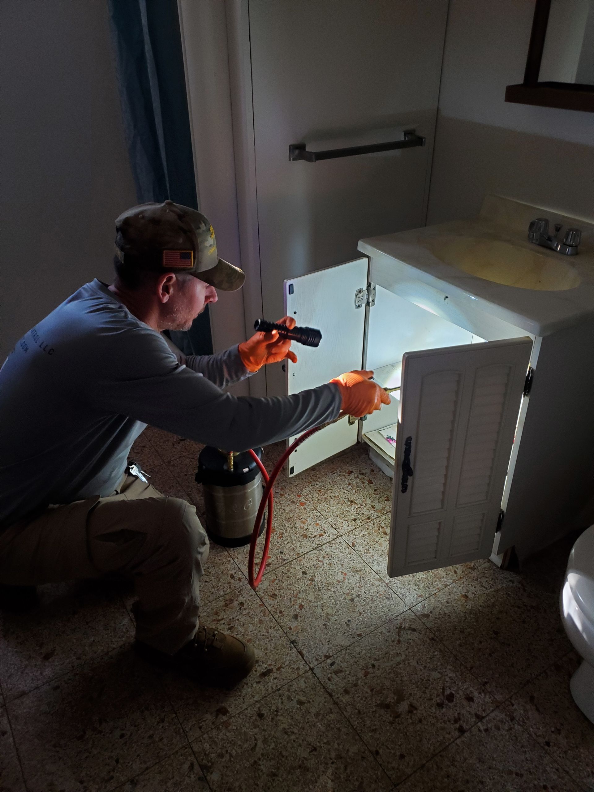 Person kneeling to use a flashlight inside an open cabinet under a sink in a dim bathroom.