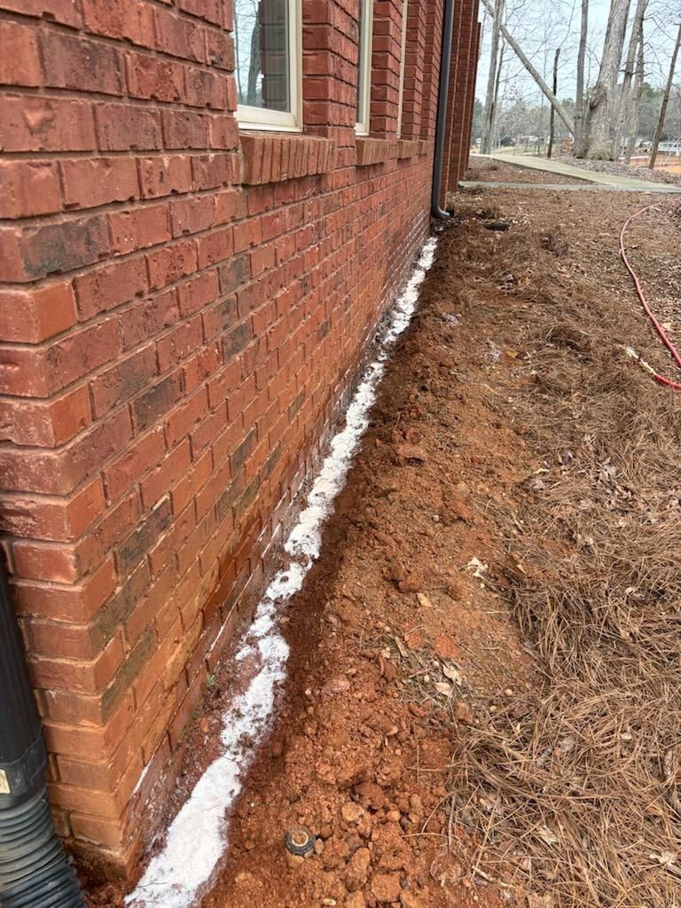 Brick house wall with a white strip of snow along the foundation beside bare soil and trees