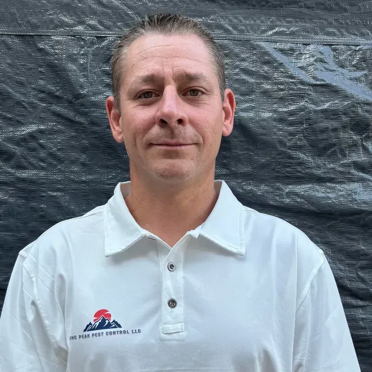 Man in white polo shirt, smiling, with logo, in front of dark textured backdrop.
