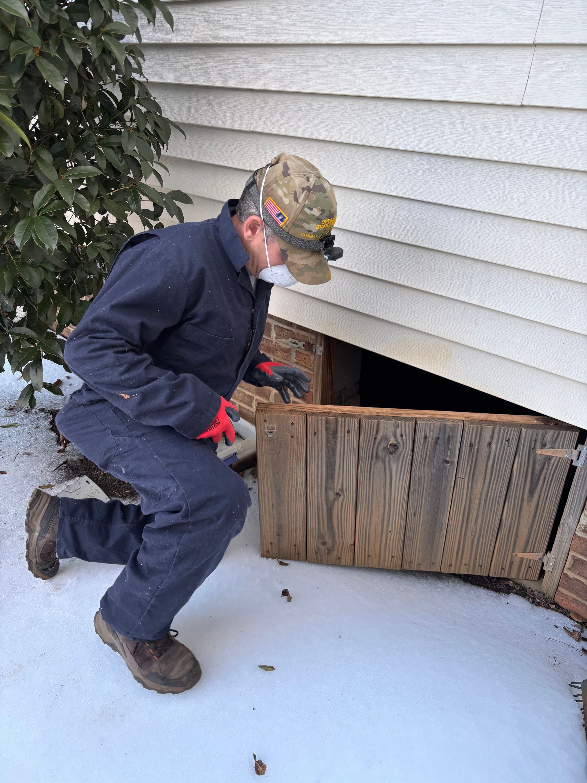 Person kneeling by an open basement hatch in snowy yard, working near the house foundation