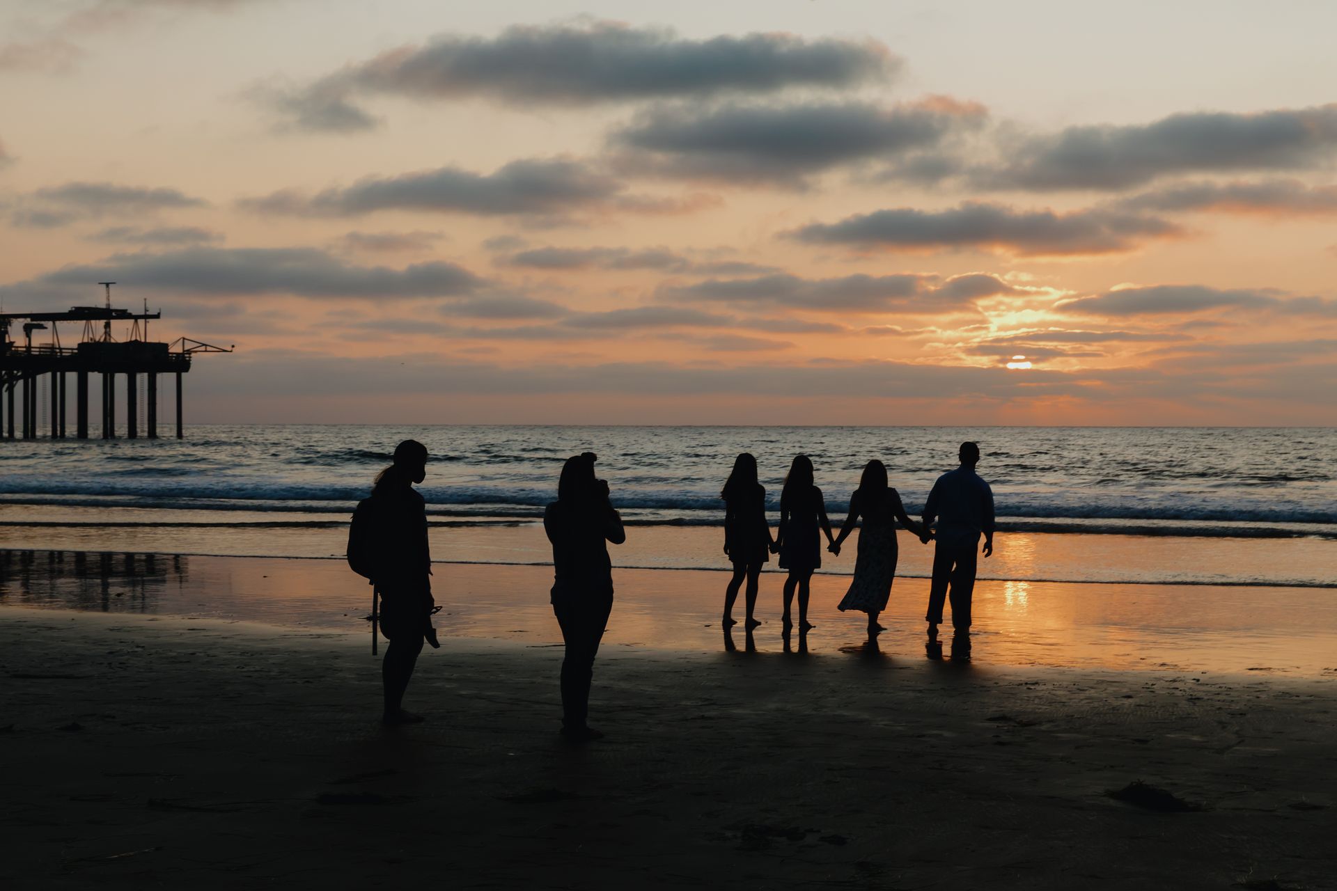 Silhouetted people walking on a beach at sunset near a pier, with orange clouds reflecting on wet sand Silhouetted people walking on a beach at sunset near a pier, with orange clouds reflecting on wet sand
