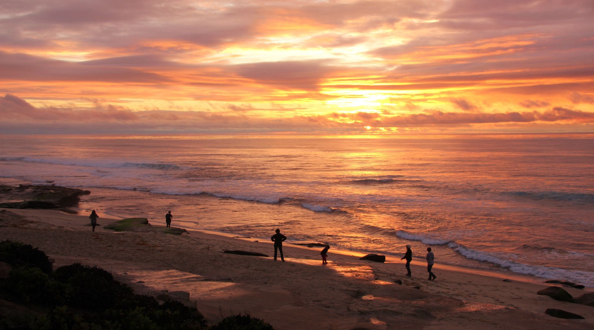 Sunset over a beach with waves, orange sky, and silhouetted people standing along the shore Sunset over a beach with waves, orange sky, and silhouetted people standing along the shore