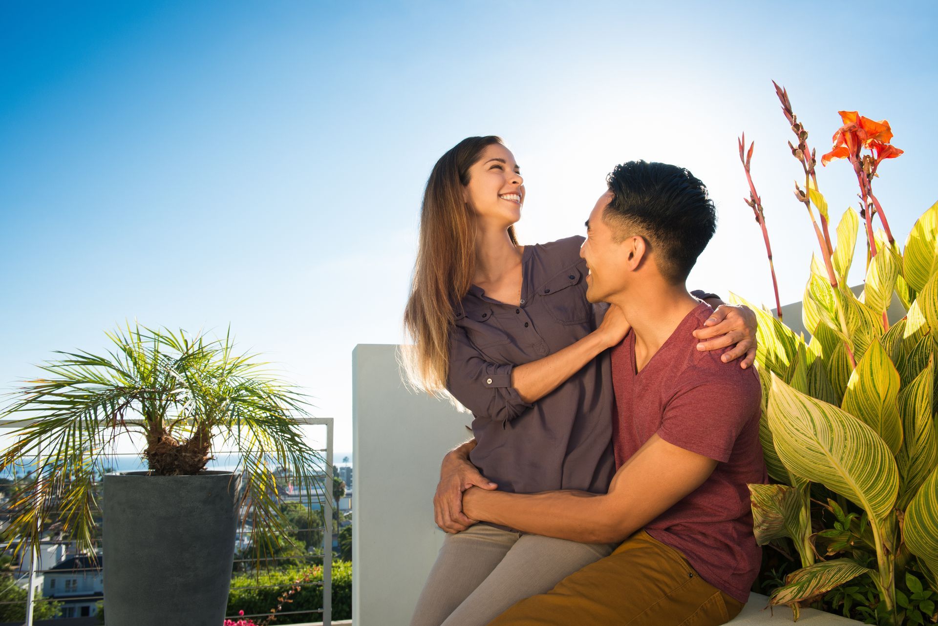 Two people sitting together on a rooftop garden, smiling and embracing beside tall plants. Two people sitting together on a rooftop garden, smiling and embracing beside tall plants.