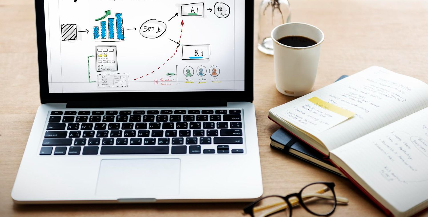 Laptop on desk showing charts, with coffee mug, open notebook, and eyeglasses nearby