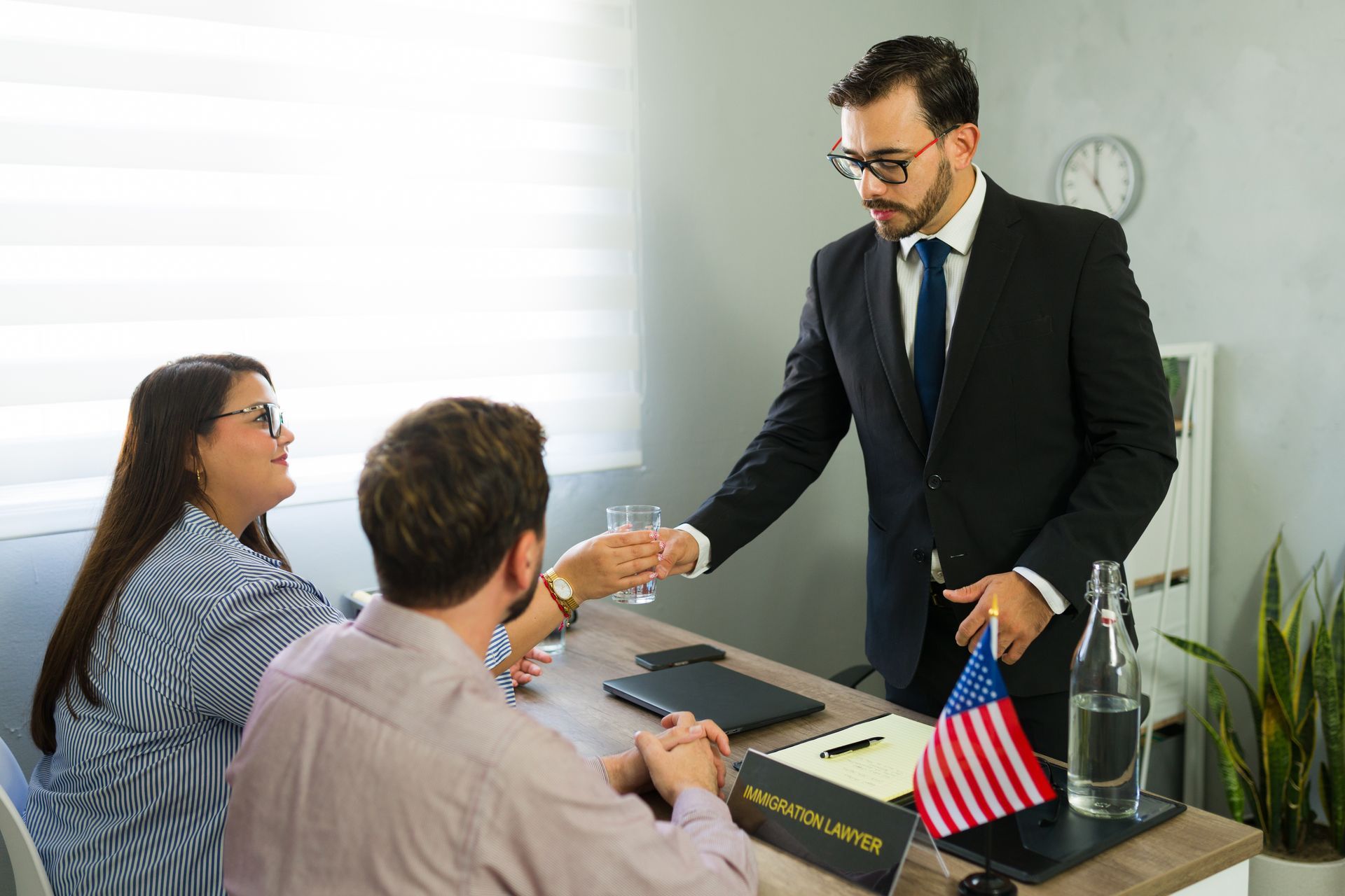 An immigration lawyer meets clients in an office with a flag and clock visible. An immigration lawyer meets clients in an office with a flag and clock visible.