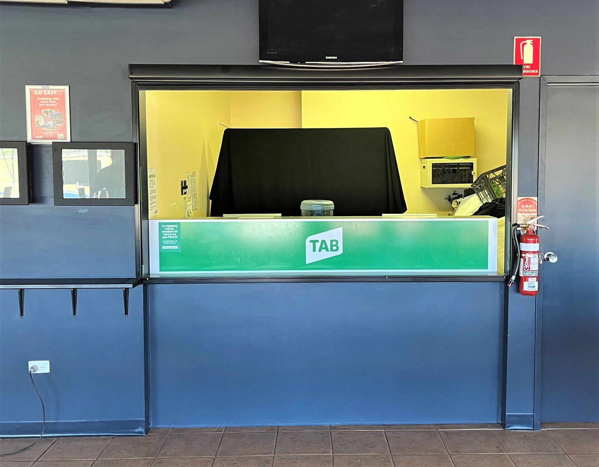 Tab Betting Window, Green Sign Blue Wall, Tv Above and Roller Door Open — Alice Mobile Blinds in Ciccone, NT