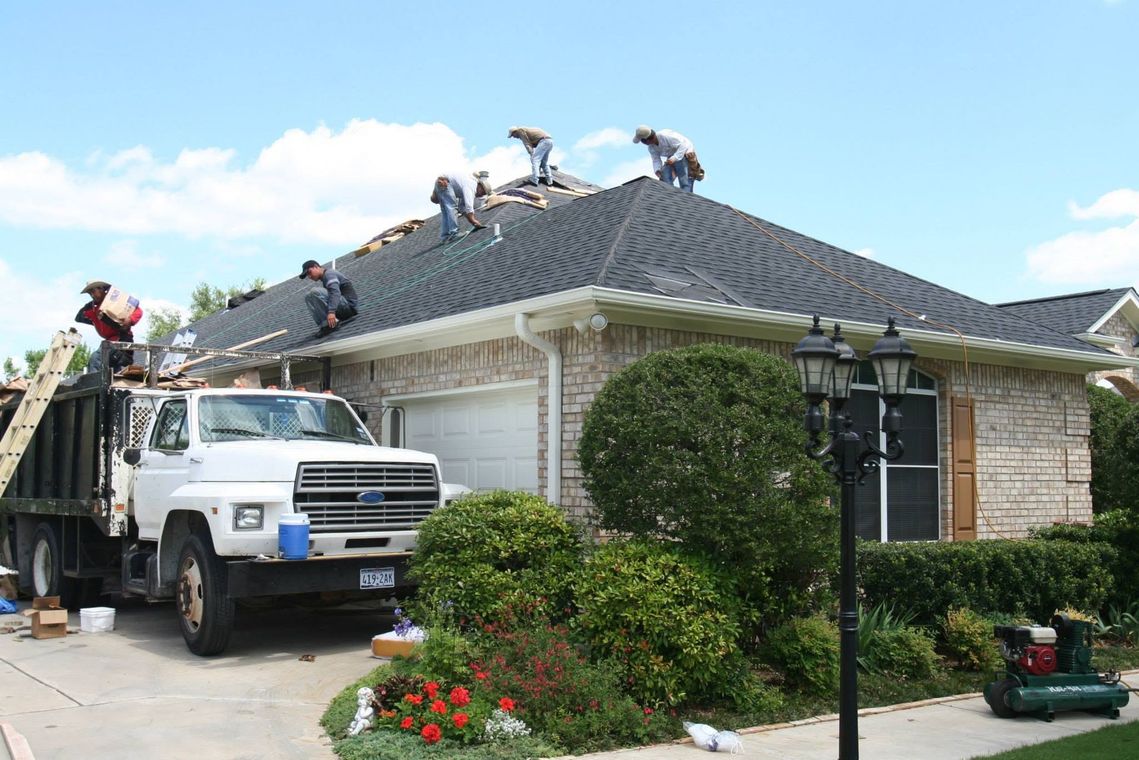 Workers on a house roof near a white truck, with shingles being repaired in a suburban neighborhood.