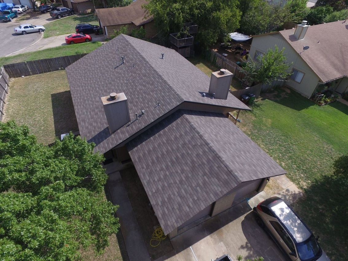 Aerial view of a gray-roofed suburban house with a driveway and parked car, surrounded by trees and lawns.