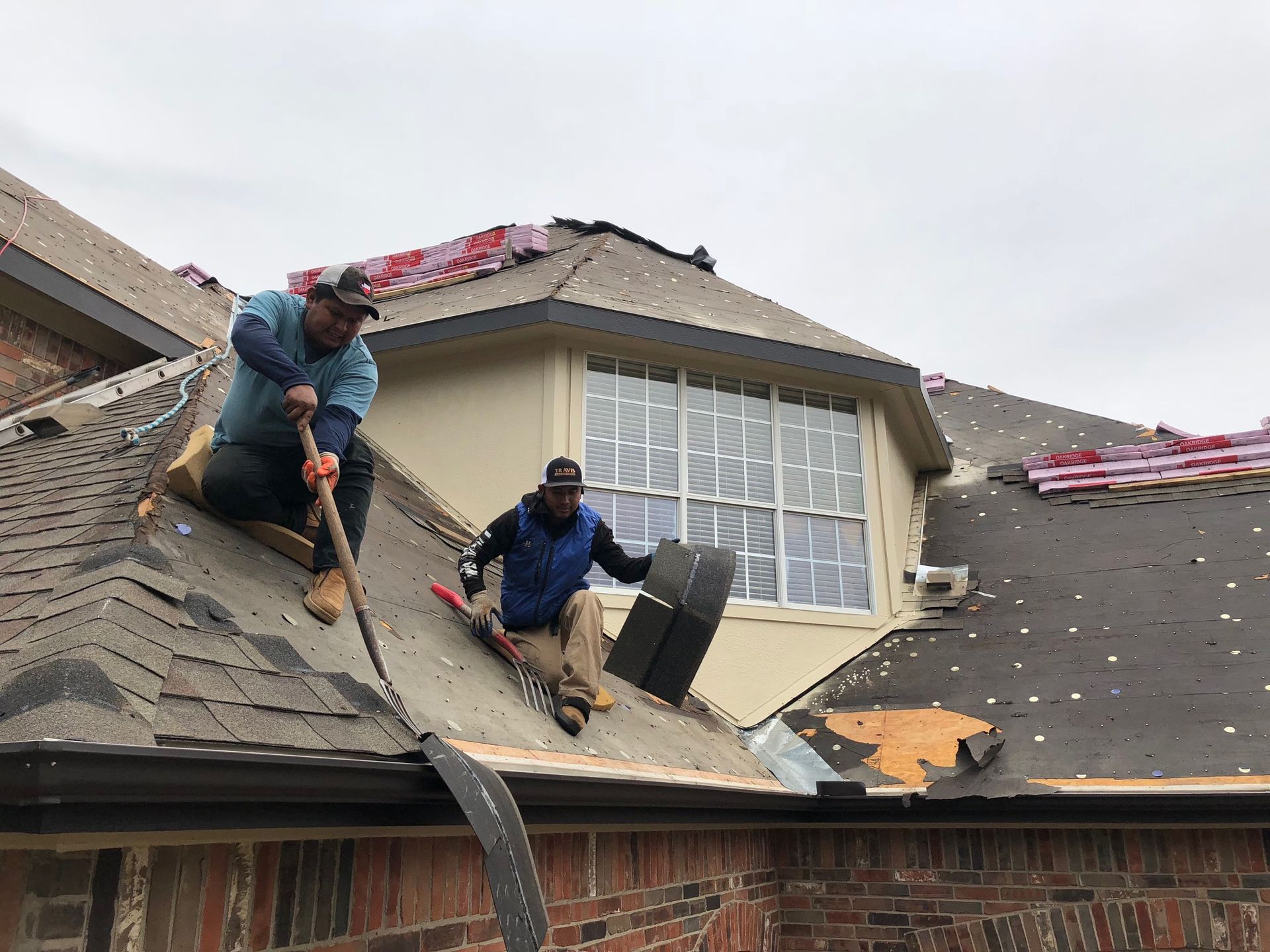 Two workers repairing a roof around a dormer window with debris and tools visible