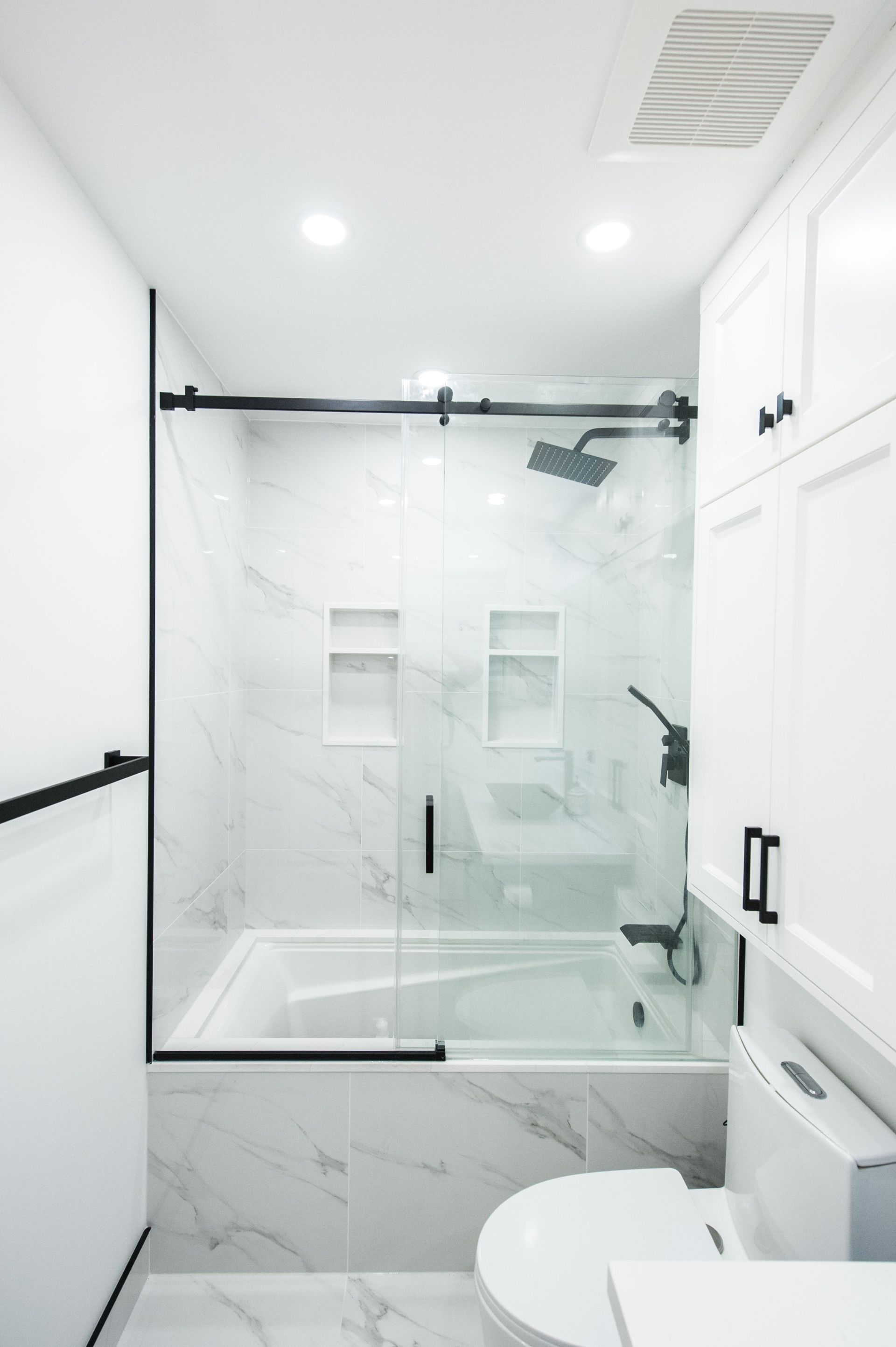Bright white bathroom with glass shower enclosure, marble tile, toilet, and vanity cabinets