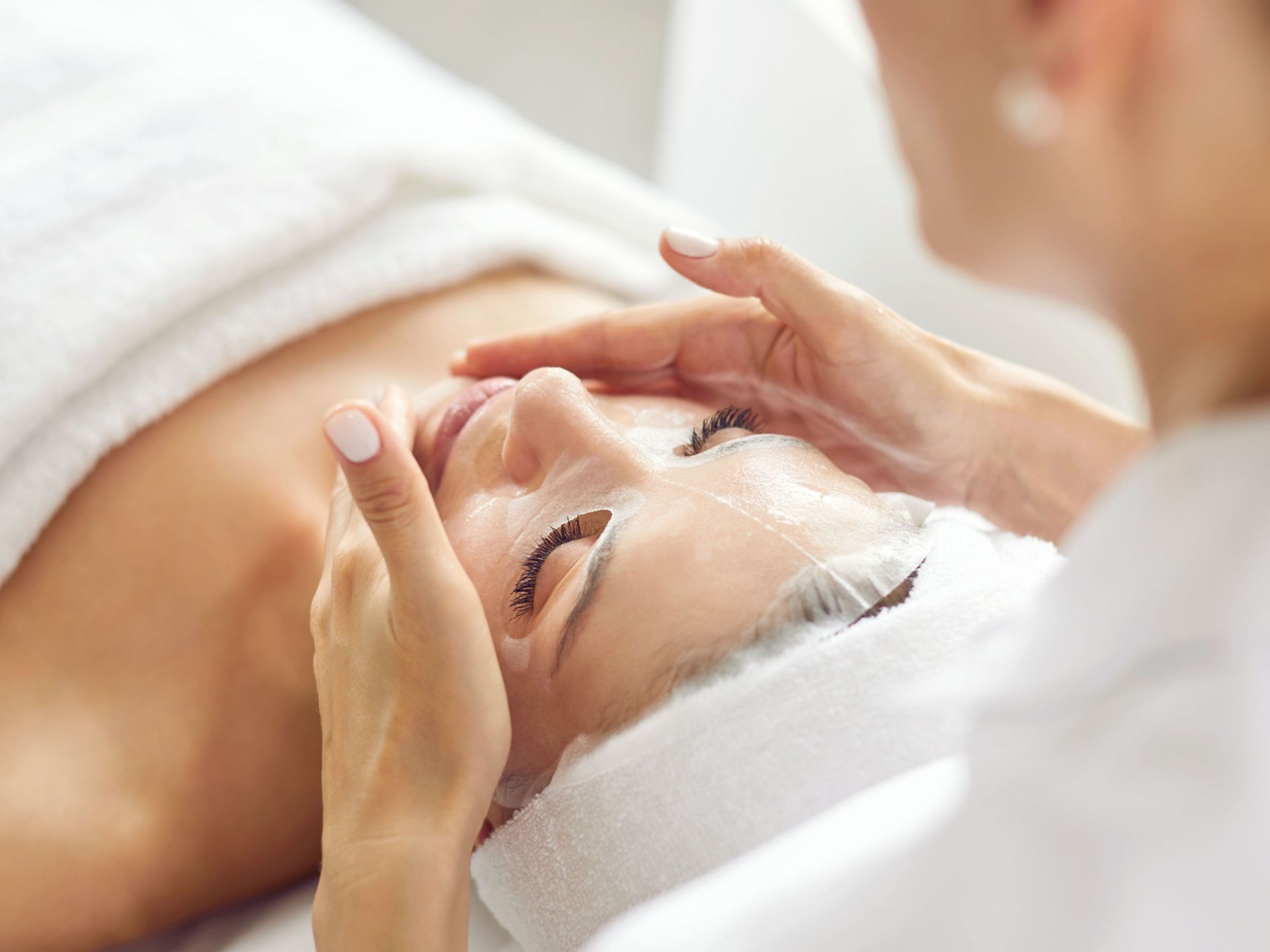 Spa facial massage with a woman lying on a treatment table, eyes closed, wrapped in white towels.