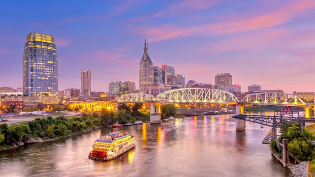 City skyline at dusk over a river with lit bridges and a boat, reflecting pink and purple sunset colors