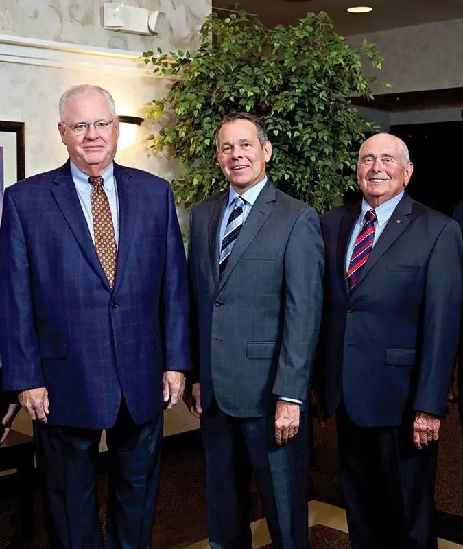 Three men in suits standing indoors, posing in front of a leafy plant.