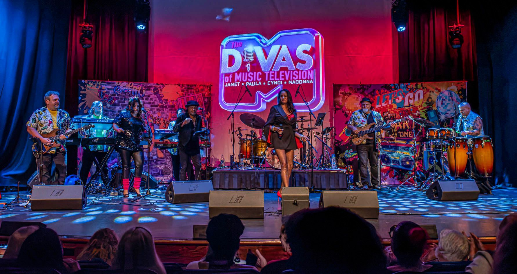 Live band performing on a colorful stage with drums, congas, and a “Divas” sign in the background.